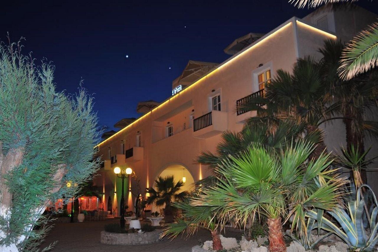 Een hotel met een witte gevel, meerdere balkons en een goed verlicht entreegebied met palmbomen en een fontein.