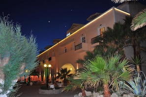 A hotel with a white facade, multiple balconies, and a well-lit entrance area with palm trees and a fountain. - Portiani Hotel (Milos)