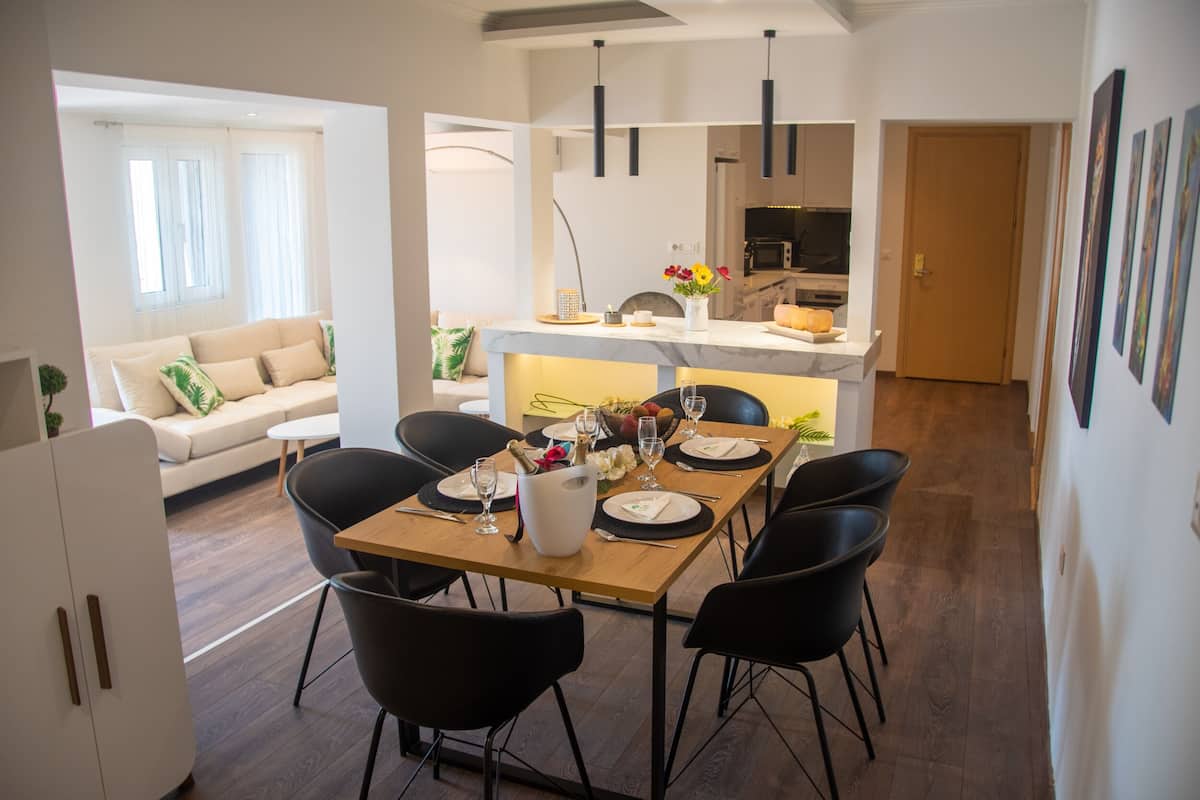 Un moderno comedor con una mesa de madera dispuesta para cuatro personas, rodeada de sillas negras. Se ve una cocina al fondo, con un microondas y un fregadero.