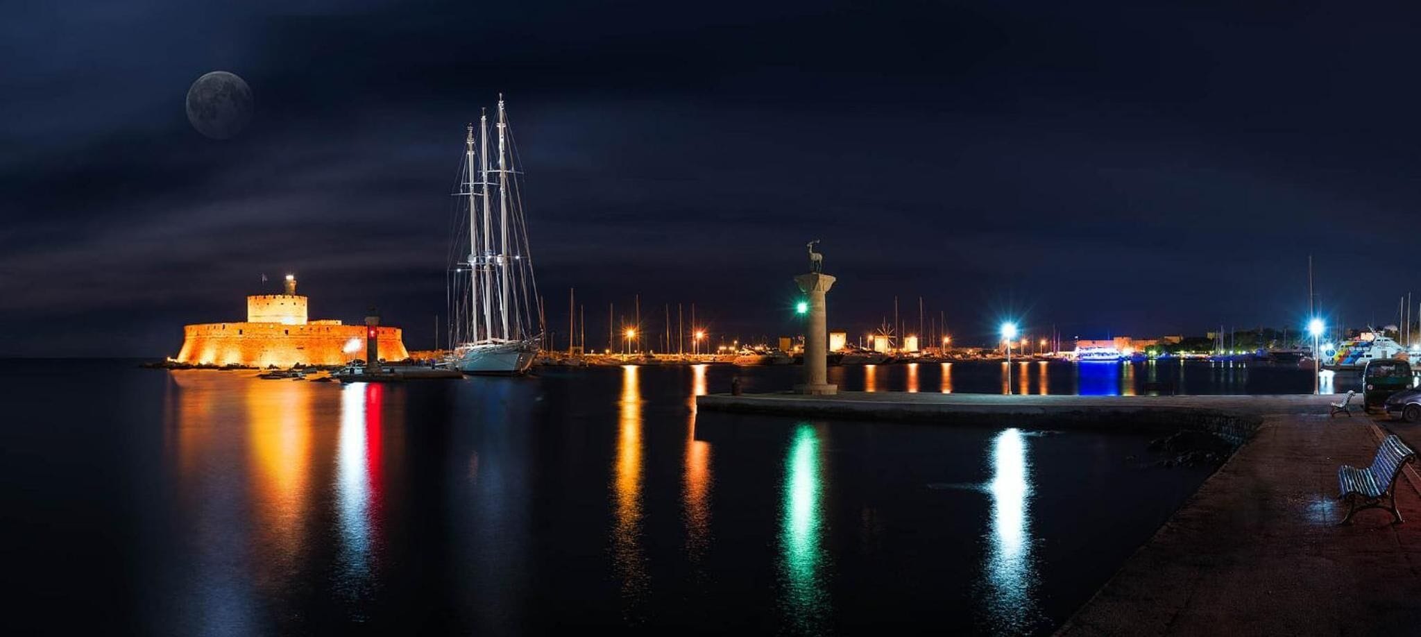 Une scène côtière avec un fort historique, un grand voilier et un phare illuminé la nuit.