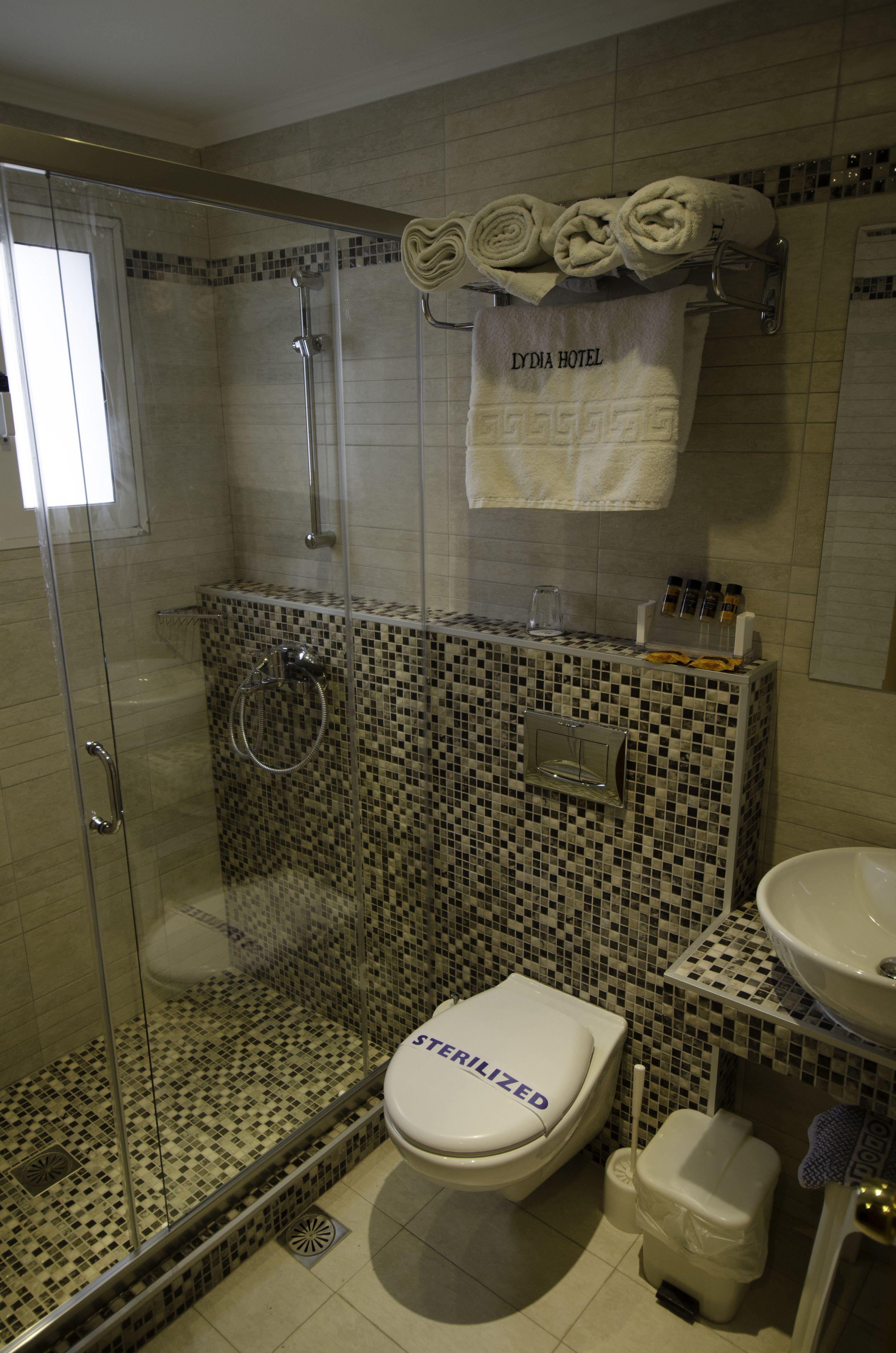 A bathroom with a glass-enclosed shower, a toilet with a "sterilized" label, a sink, and a towel rack with folded towels.
