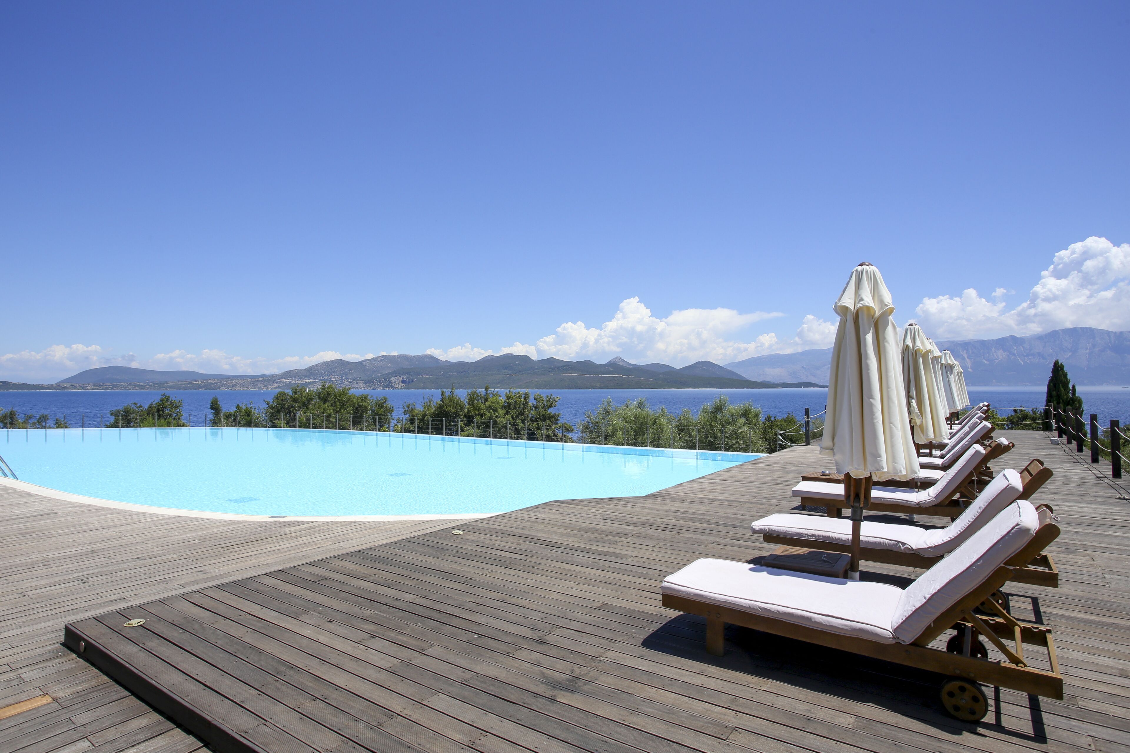 A wooden deck with a pool, lounge chairs, and umbrellas overlooking a body of water with mountains in the background.