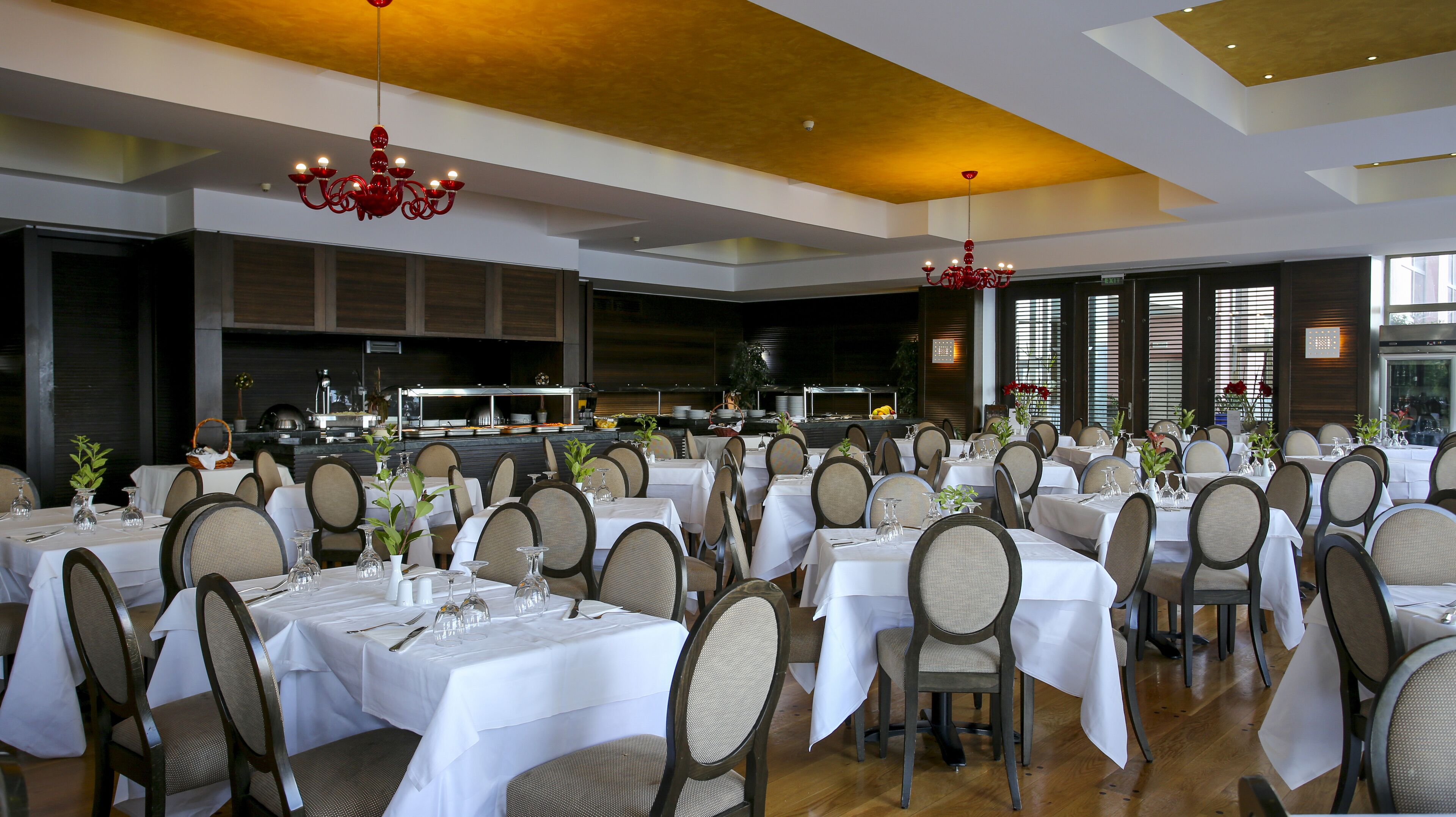 A spacious dining area with multiple tables set for guests, featuring a modern chandelier and a view of the kitchen in the background.