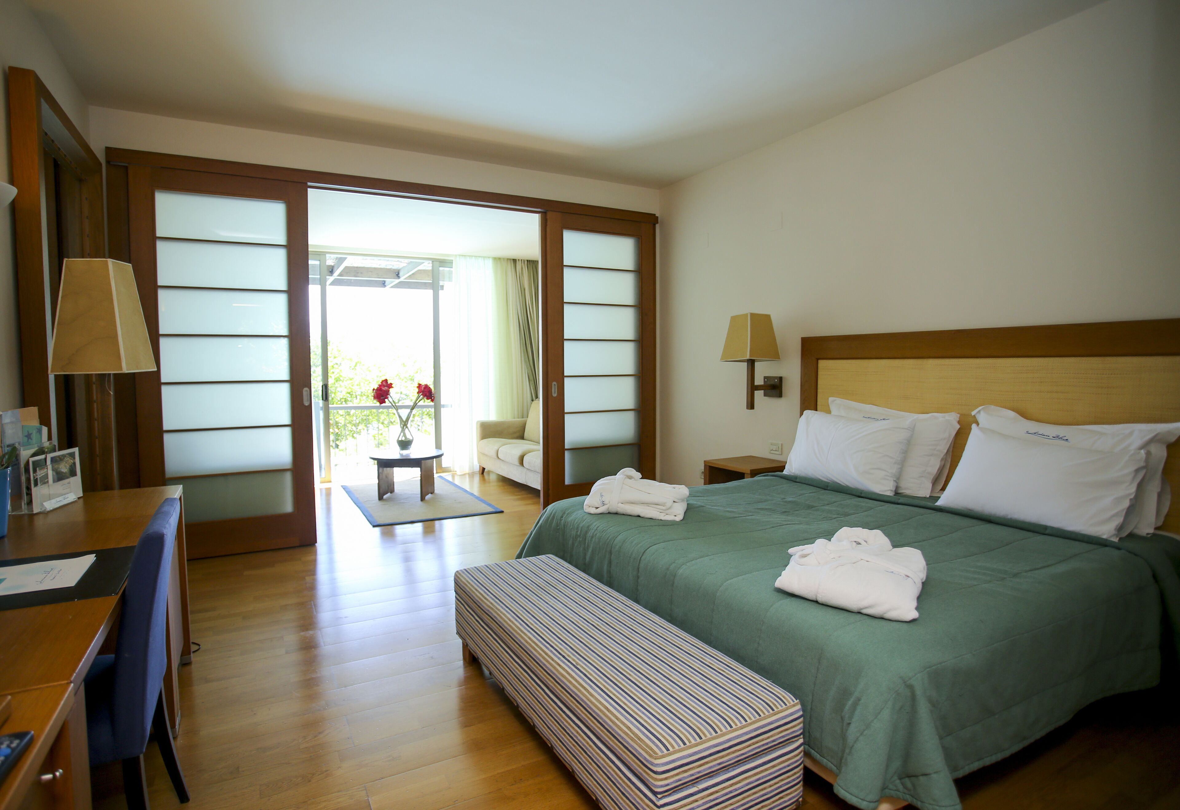 A hotel room with a bed, a desk, a chair, a bench, and a view of the outdoors.