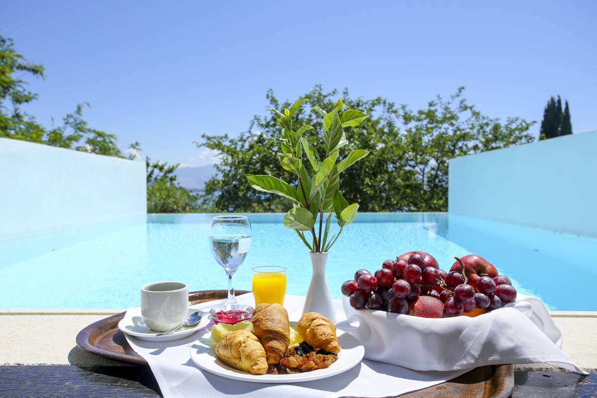 A breakfast tray with croissants, fruit, and juice by a poolside with a clear view of the pool and trees in the background.