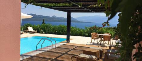 Un área junto a la piscina con una terraza de madera, sillas de descanso y una pérgola cubierta de plantas trepadoras.