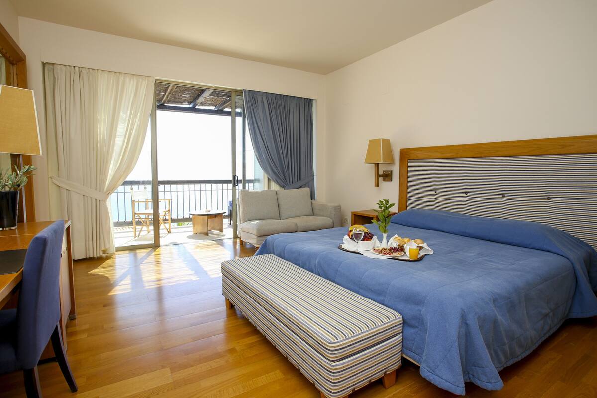 A hotel room with a bed, a desk, a chair, a bench, a sofa, a table, and a window with curtains.