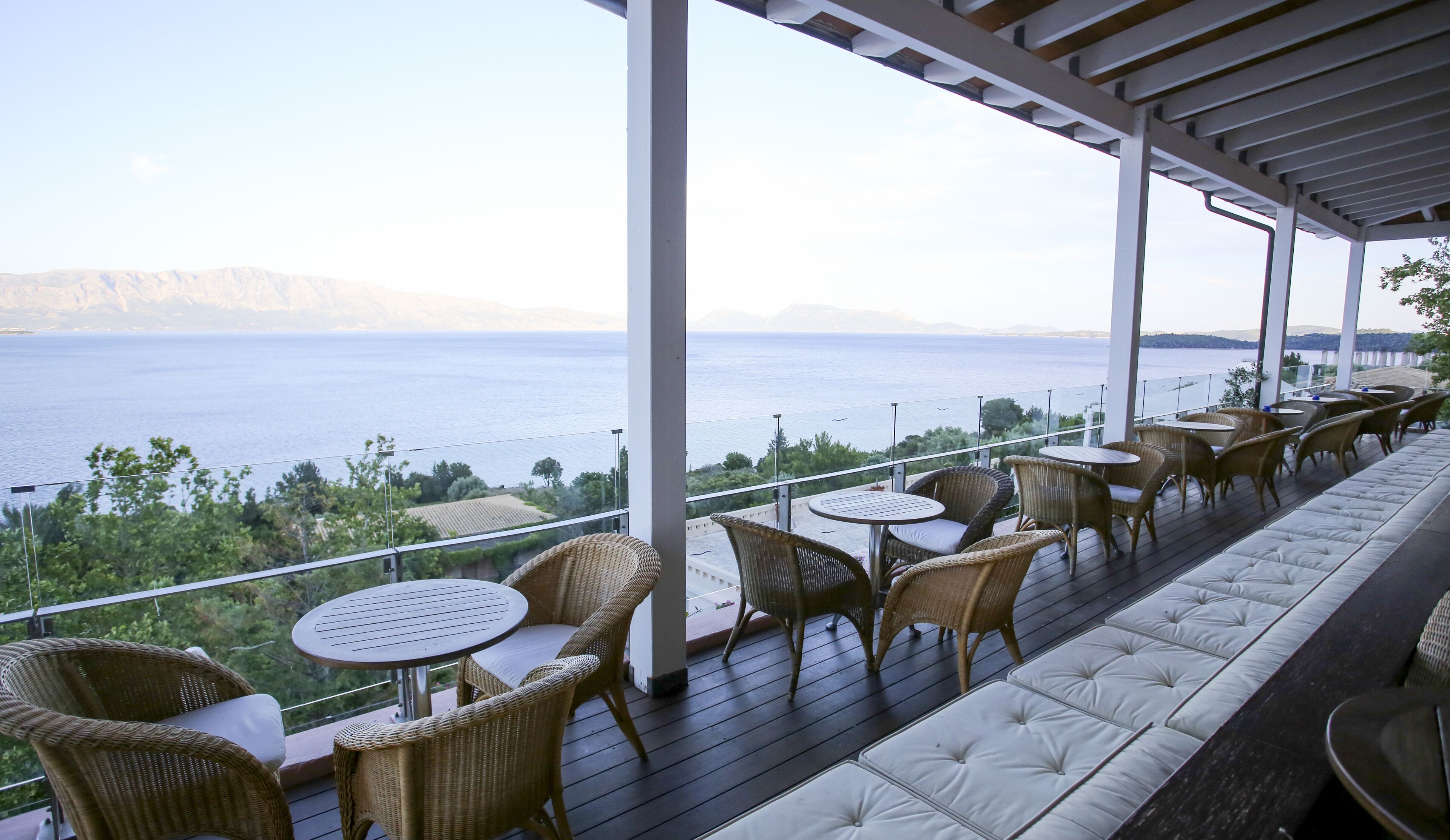 A covered outdoor seating area with wicker furniture and a cushioned bench, overlooking a body of water and distant mountains.