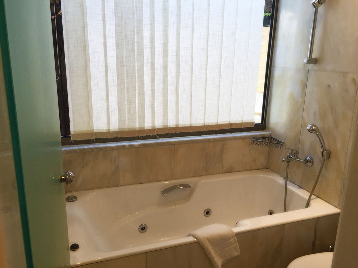 A bathroom with a bathtub, a window with blinds, a shower, and a toilet.
