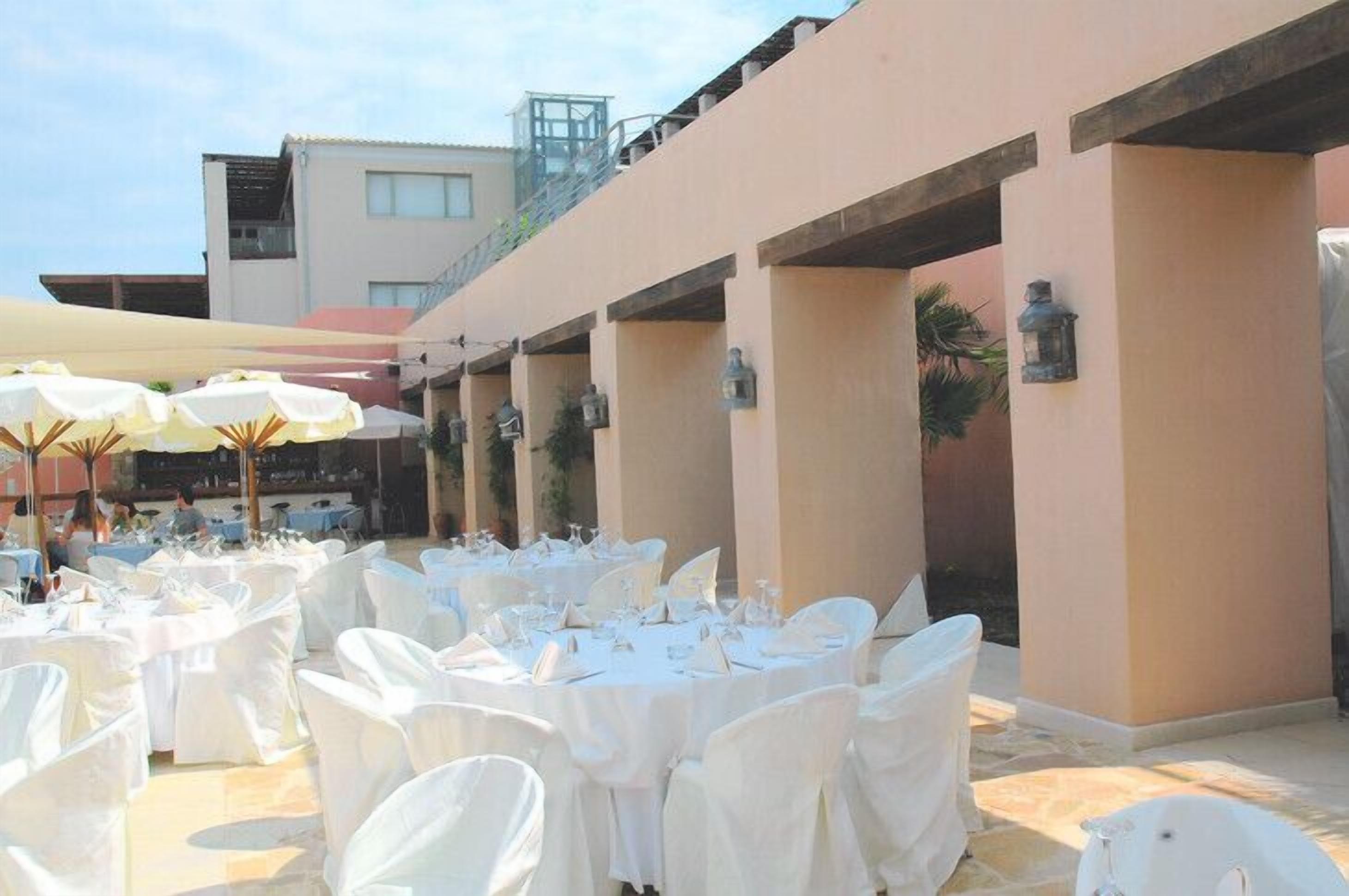 Un área de comedor al aire libre con mesas redondas dispuestas para comensales, cubiertas con manteles blancos y sillas, rodeada de paredes beige con faroles.