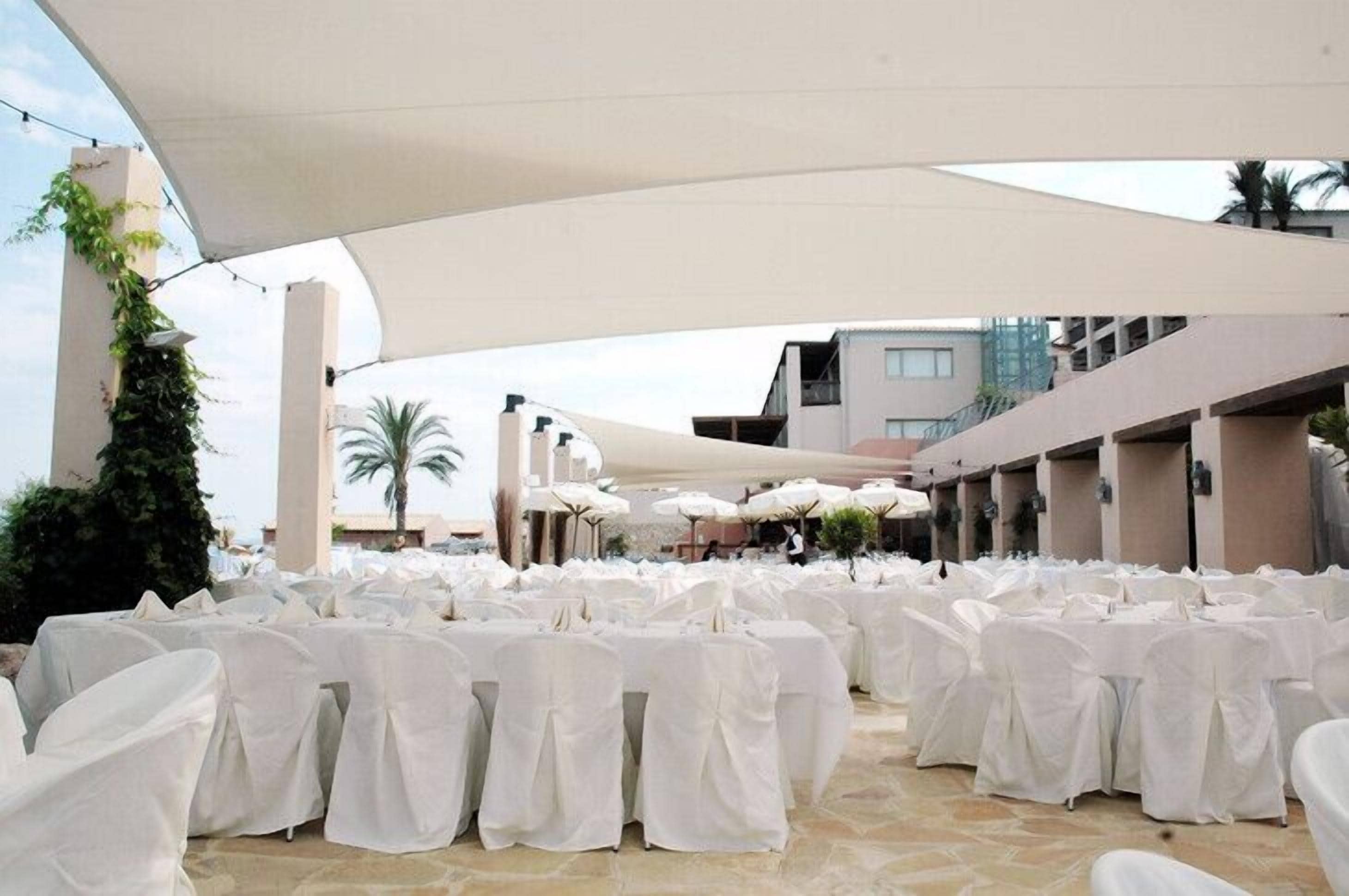 An outdoor seating area with white-covered tables and chairs, shaded by large white canopies, set against a backdrop of buildings and palm trees.