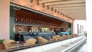 A covered outdoor seating area with cushioned chairs and tables, a bar in the background, and a wooden ceiling structure. - Ionian Blue Bungalows And Spa Resort (Lefkada)