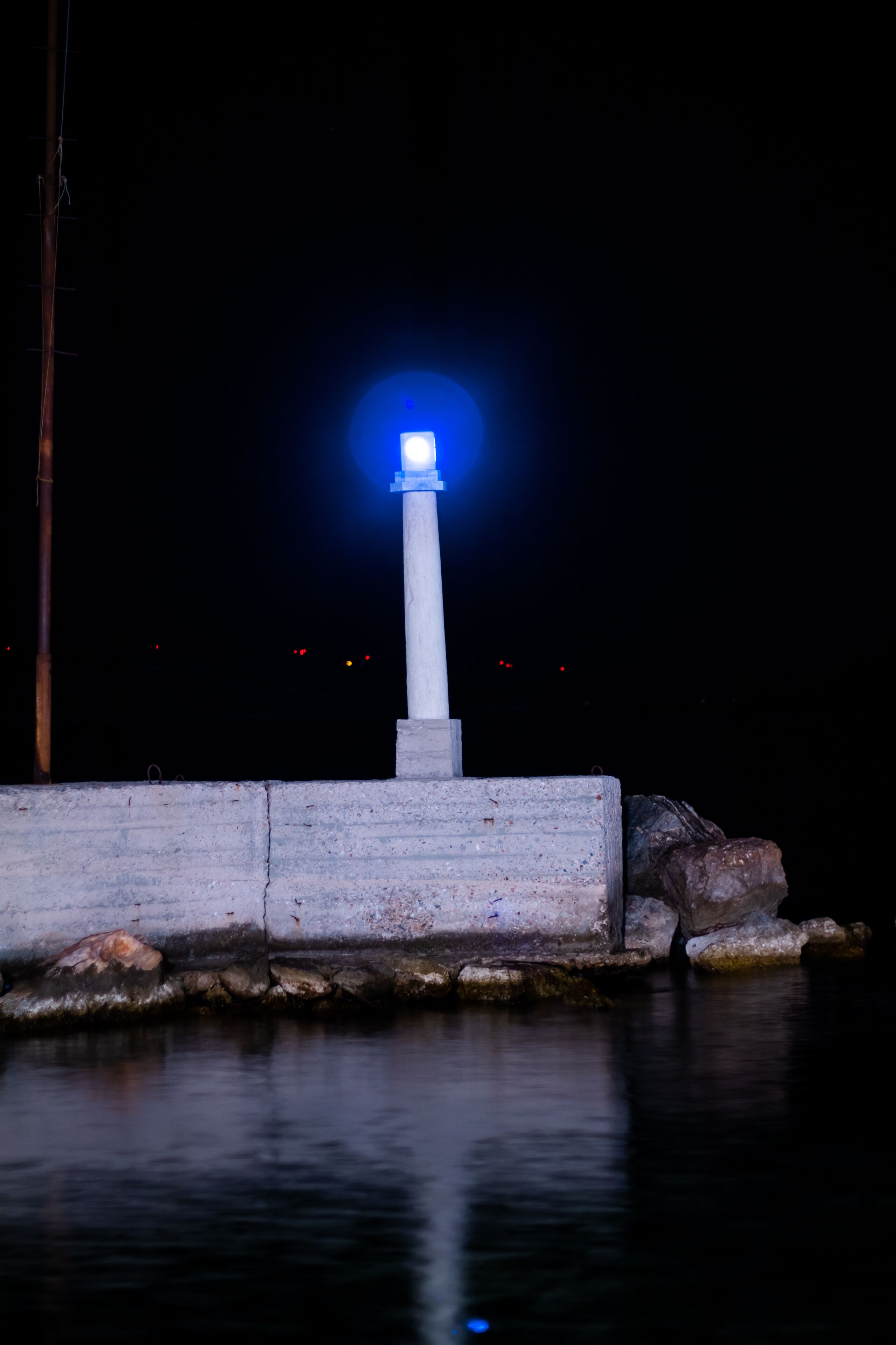 Un faro de noche con una luz azul brillante en la parte superior, situado junto a una barrera de hormigón y rocas a la orilla del agua.