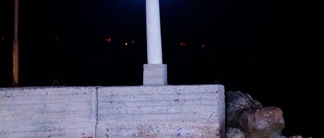 Un faro de noche con una luz azul brillante en la parte superior, situado junto a una barrera de hormigón y rocas a la orilla del agua.
