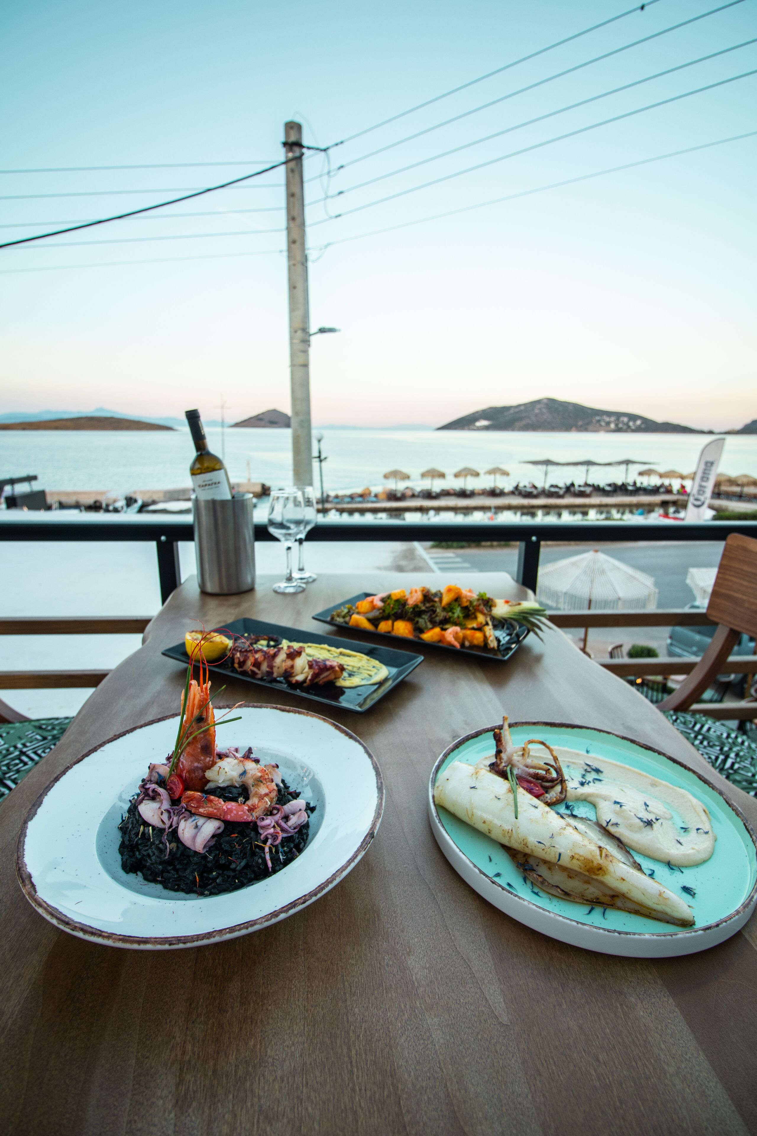 A table with seafood dishes, a wine bottle, and a glass of wine, overlooking a beach with umbrellas and people.