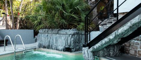 Una piscina con cascada, rodeada de plantas tropicales y un muro de piedra.