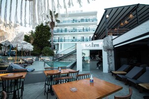 Un bar en la azotea con mesas y sillas de madera, una piscina y vistas a un edificio de varios pisos con balcones.