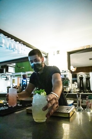 A bartender preparing a drink at a bar with a metal counter, glasses, and bottles in the background. - Kiani Akti Hotel (Markopoulo Mesogaias)