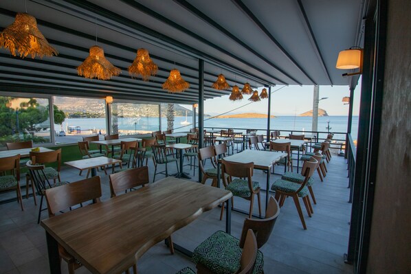 An outdoor dining area with tables and chairs, large windows, and hanging lights. - Kiani Akti Hotel (Markopoulo Mesogaias)