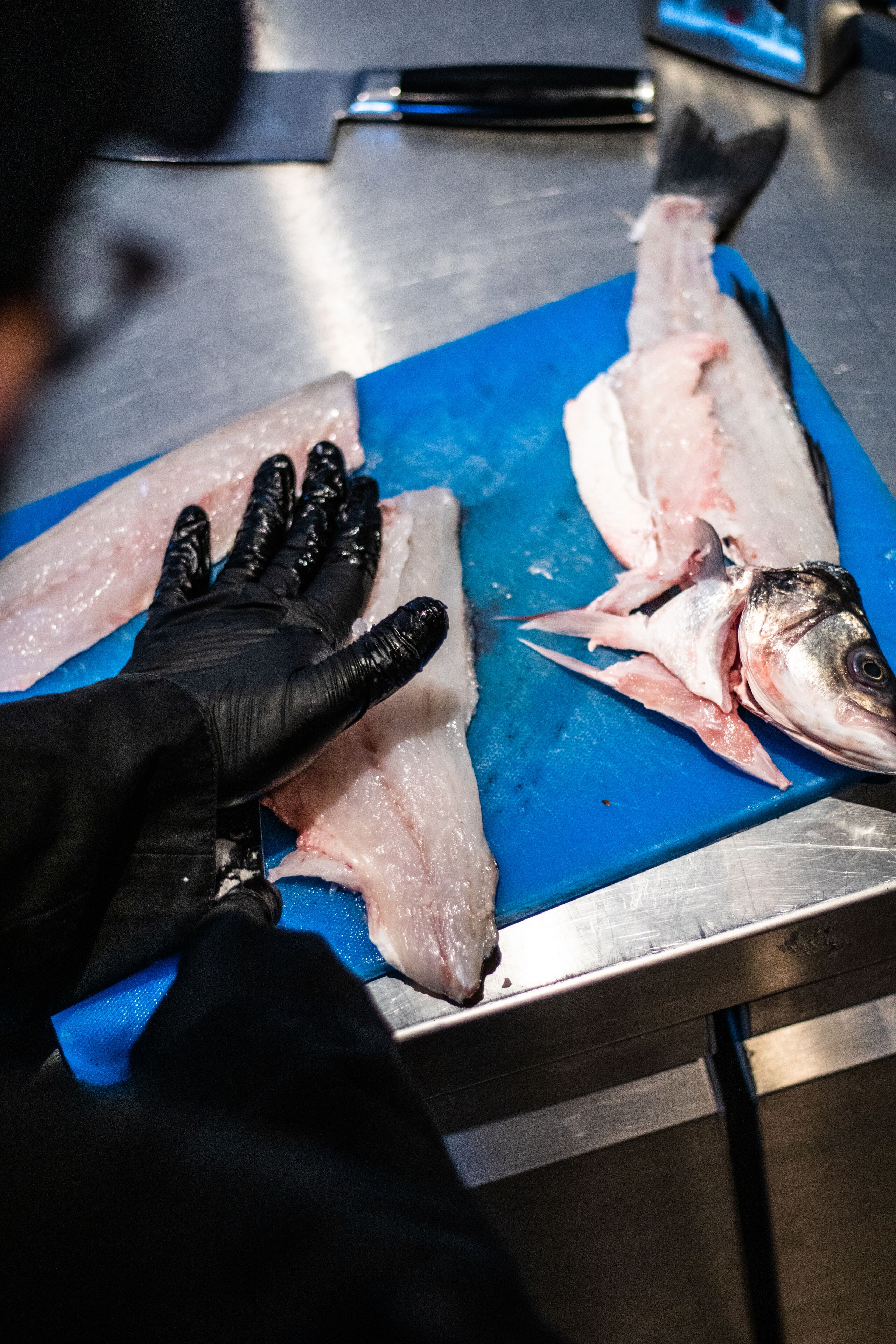 Uma pessoa com luvas pretas manuseando peixe cru numa tábua de corte azul.