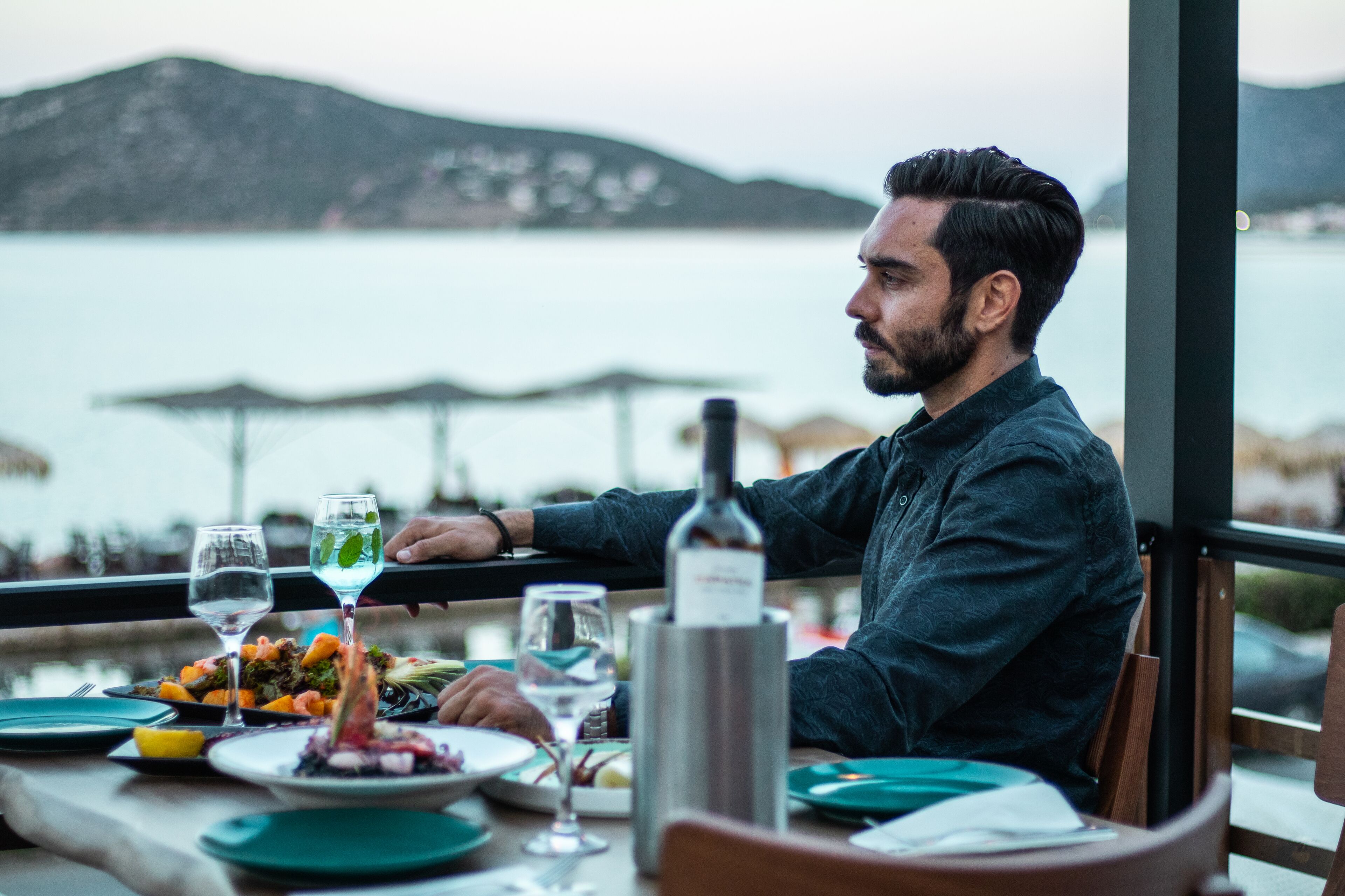 Una persona cenando en una mesa con vista al agua y montañas al fondo.