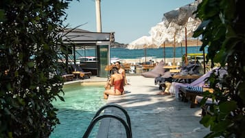 Zona junto a la piscina con sillones y sombrillas, vista despejada al mar y una construcción alta al fondo.