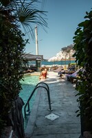 Zona junto a la piscina con sillones y sombrillas, vista despejada al mar y una construcción alta al fondo.