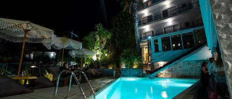 Une piscine bordée d’un mur en pierre et un escalier menant à un niveau supérieur. Des chaises longues et des parasols sont disposés autour de la piscine.