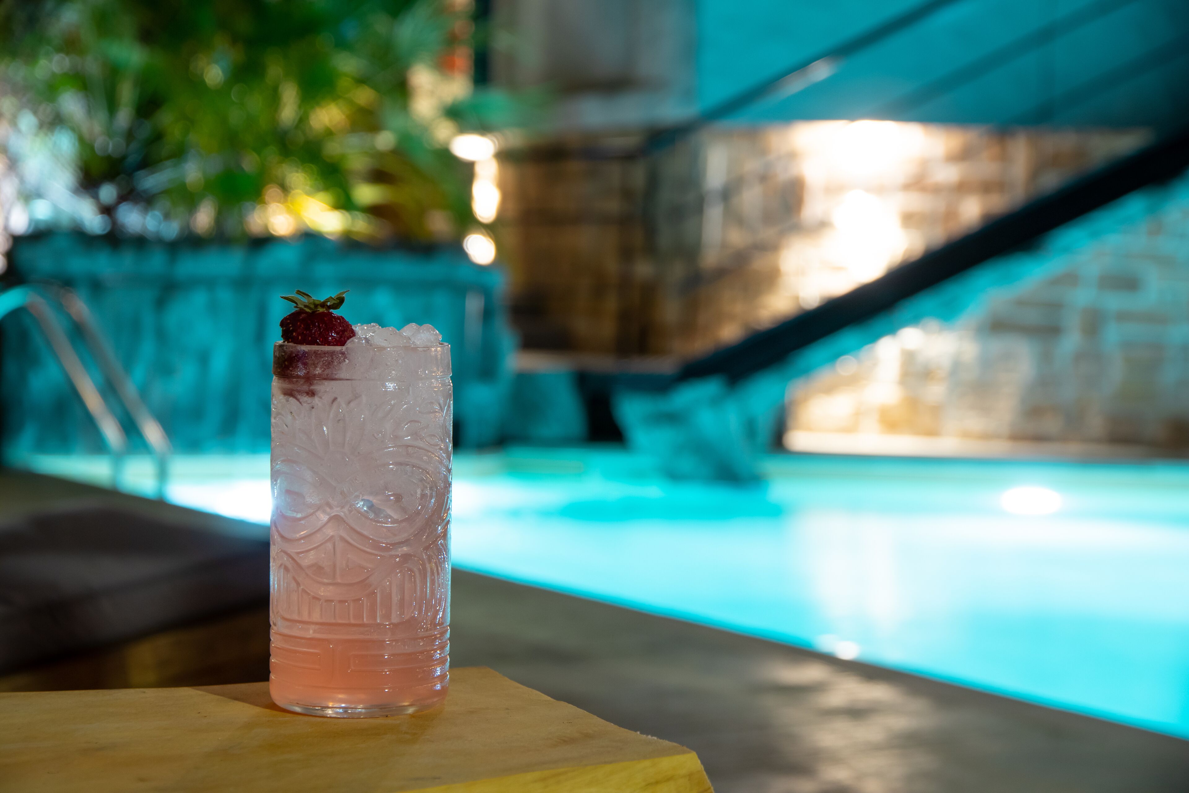 A cocktail glass with a strawberry on the rim, placed on a wooden surface near a pool.