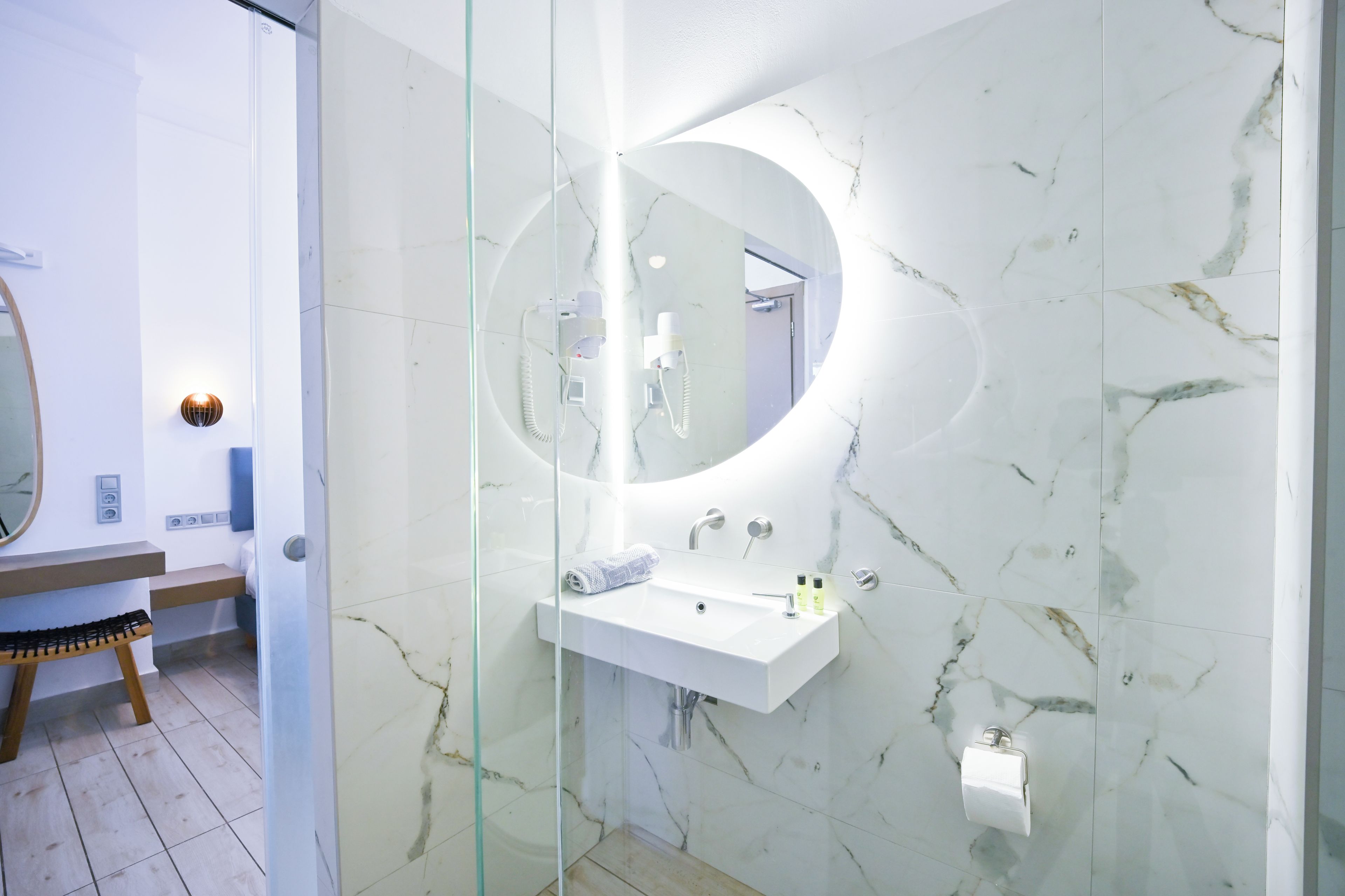 Une salle de bain moderne avec une paroi en verre, un miroir rond, un lavabo blanc et une serviette posée sur le comptoir.