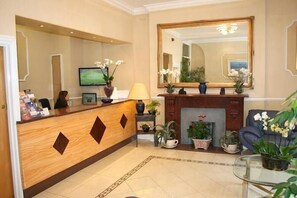 A reception area with a wooden reception desk, a television, a fireplace, and several potted plants. - Avon Hotel (London)