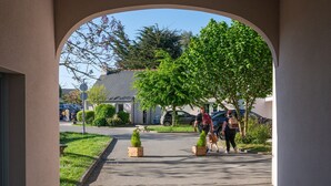 Une vue à travers une arche donnant sur une rue résidentielle, avec des maisons, des voitures garées et des personnes qui se promènent.