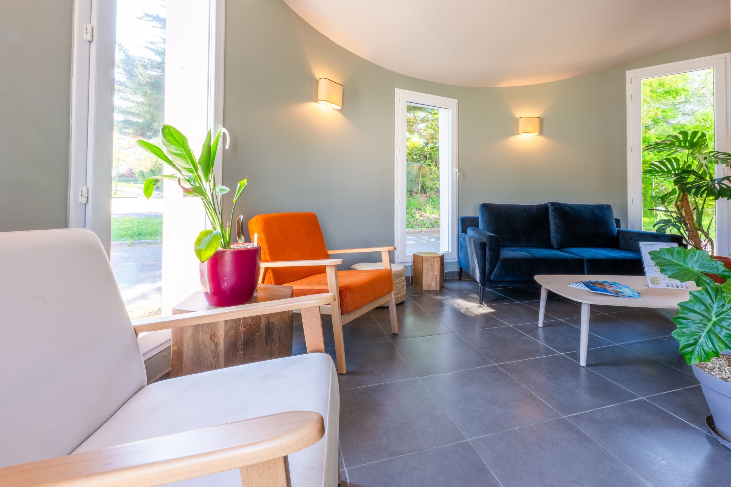 Una sala de estar moderna con un sofá blanco, una silla naranja, un sofá azul y una mesa de centro. Hay dos plantas en macetas, una sobre una mesita y otra cerca del sofá. La habitación tiene un techo curvo y ventanas amplias con vistas a la vegetación exterior.