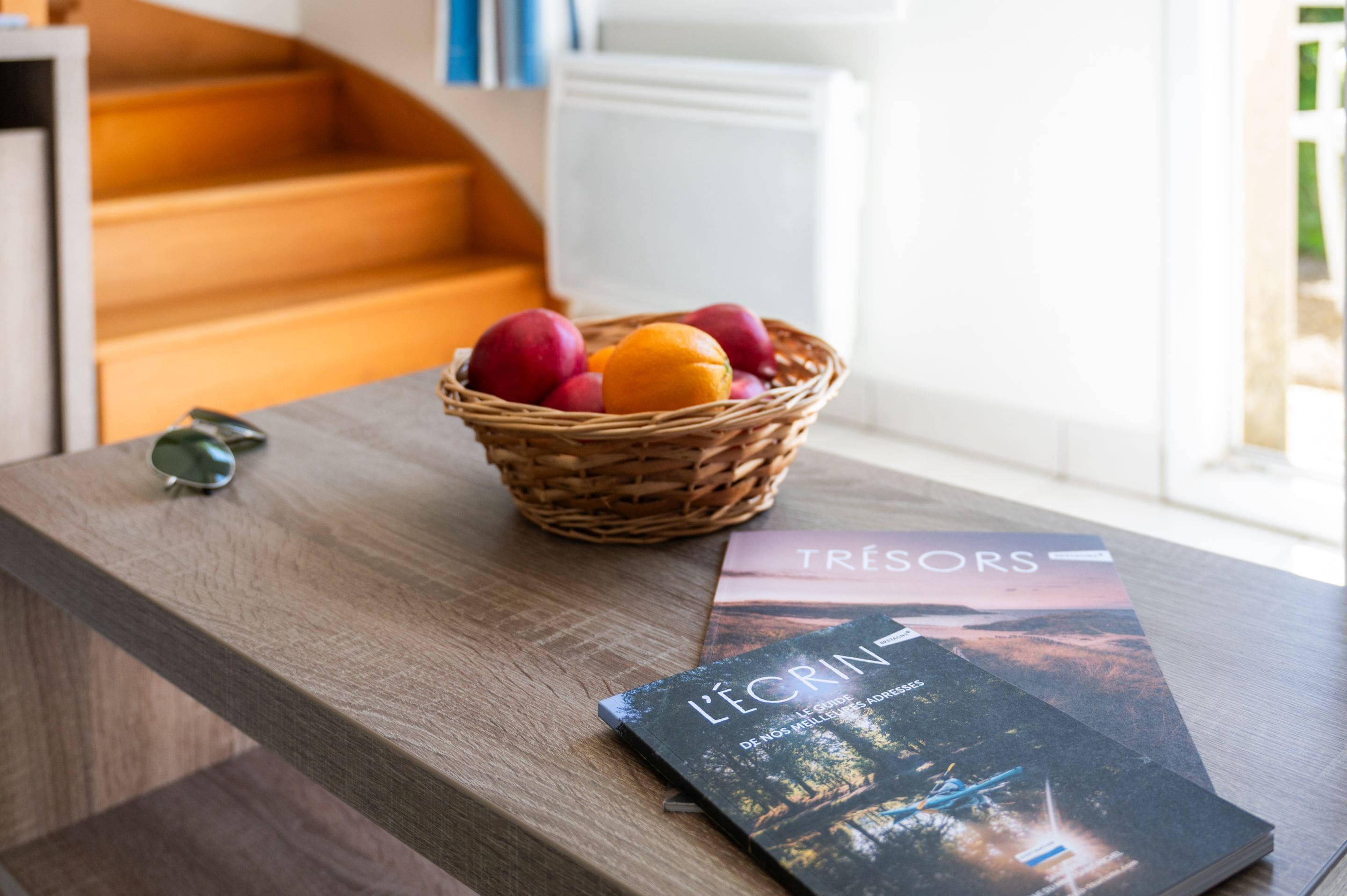 A wooden table with a wicker basket containing fruits, a pair of sunglasses, and a magazine titled "L'écrin".