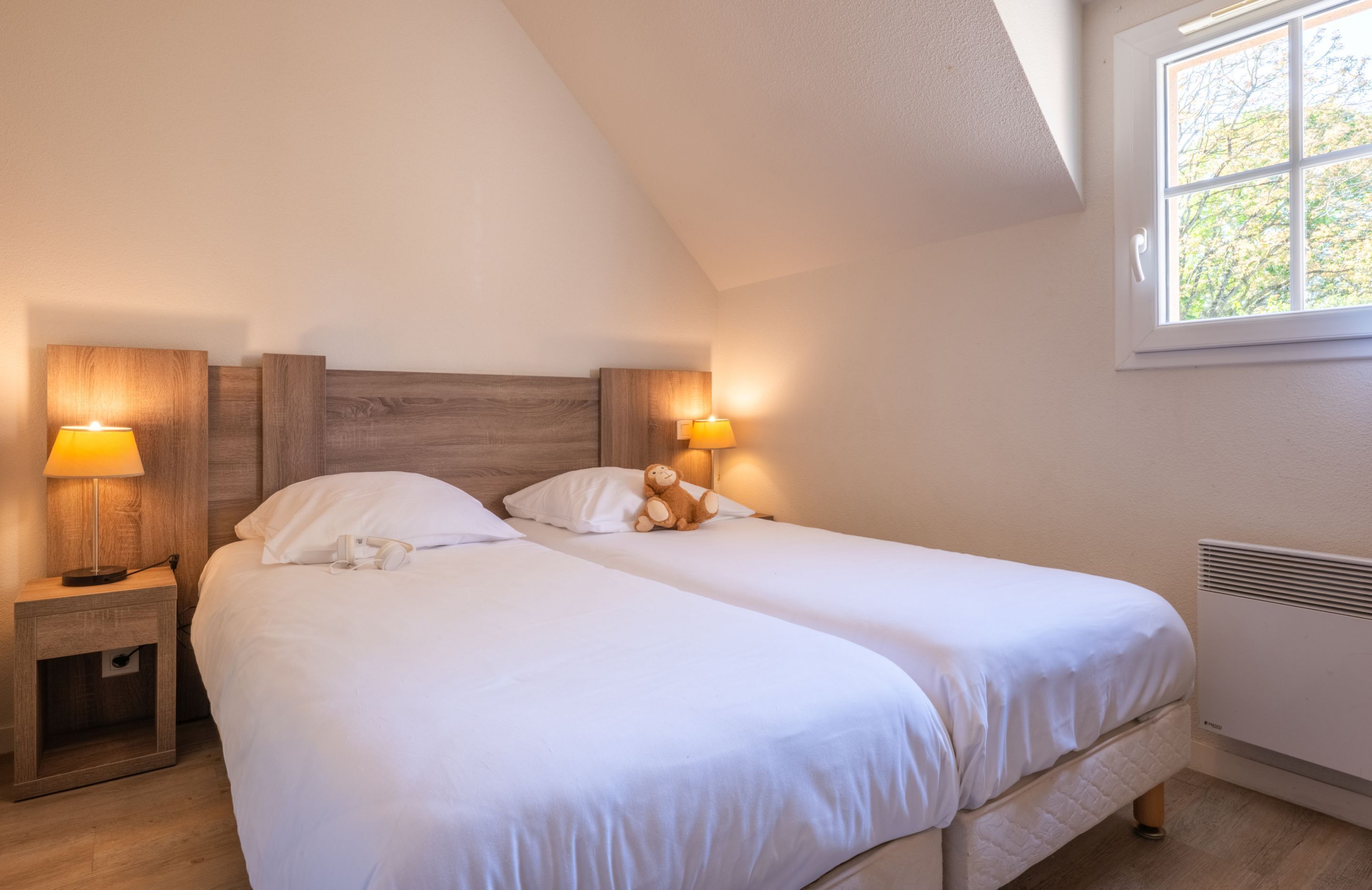 A bedroom with two beds, wooden headboards, bedside lamps, and a window with a view of trees.