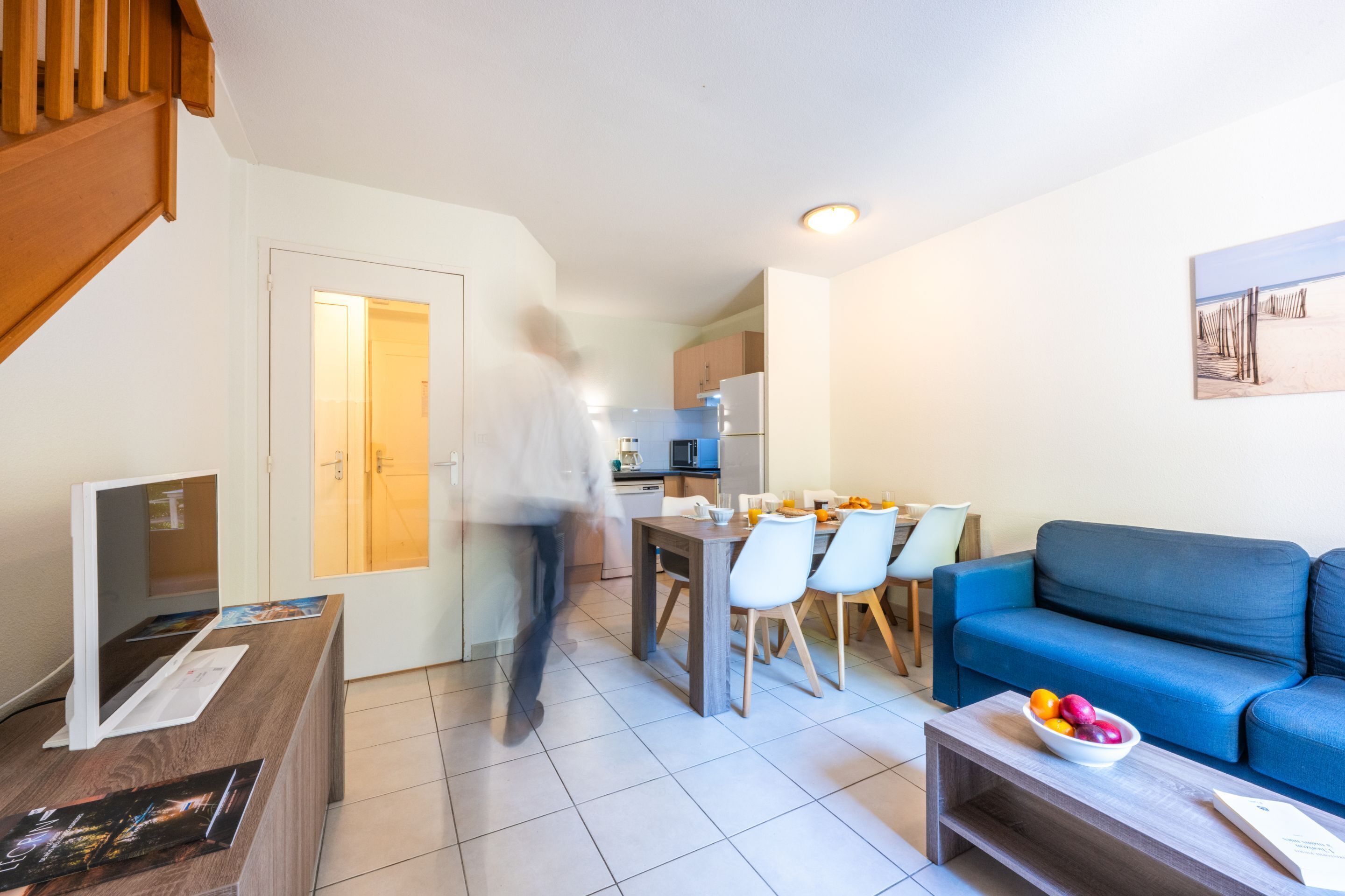 A modern living room with a blue sofa, a dining table with chairs, a kitchen in the background, and a staircase leading to an upper level.