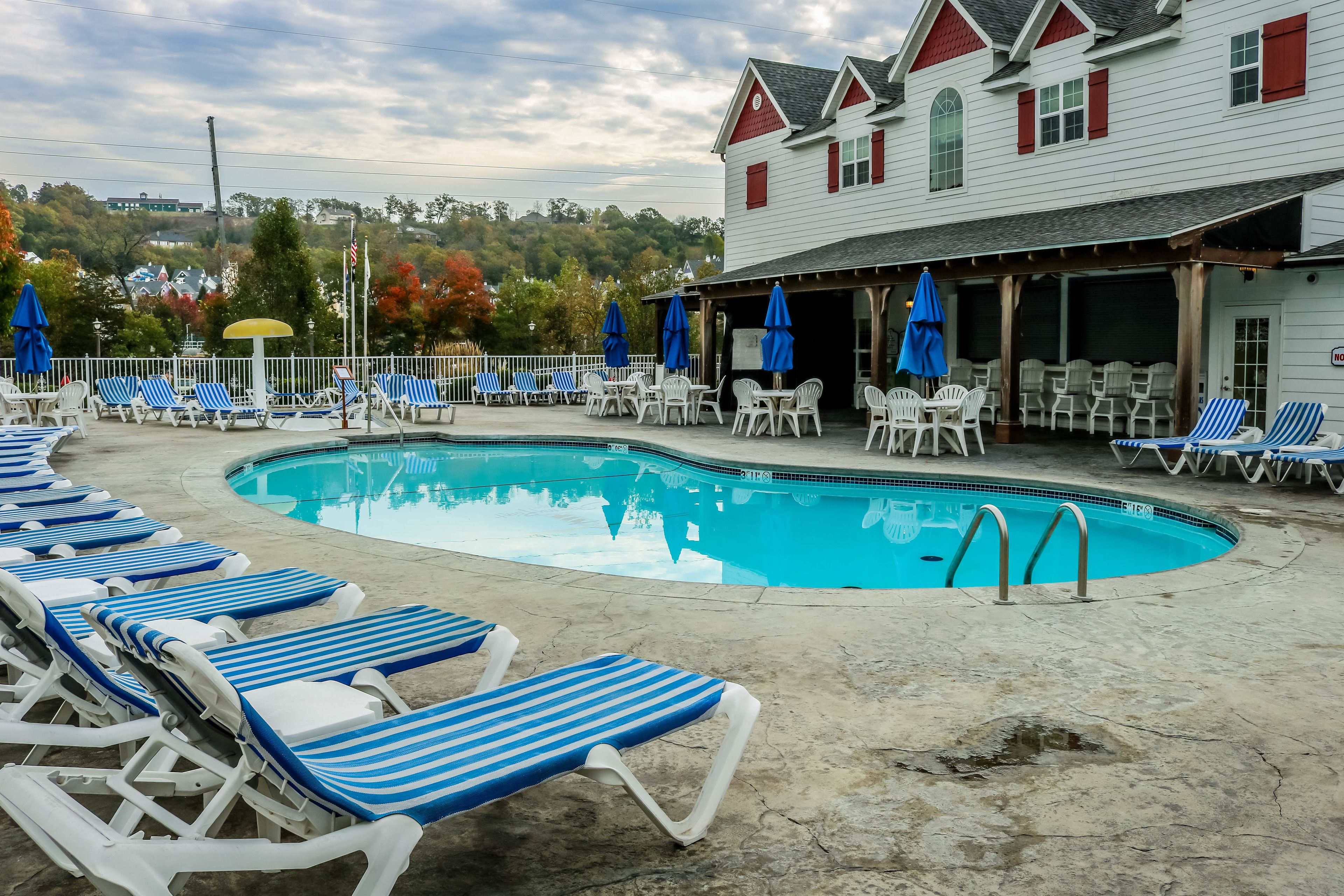 Zona de piscina con sillones de descanso y sombrillas, un área de estar cubierta y un edificio con contraventanas rojas.