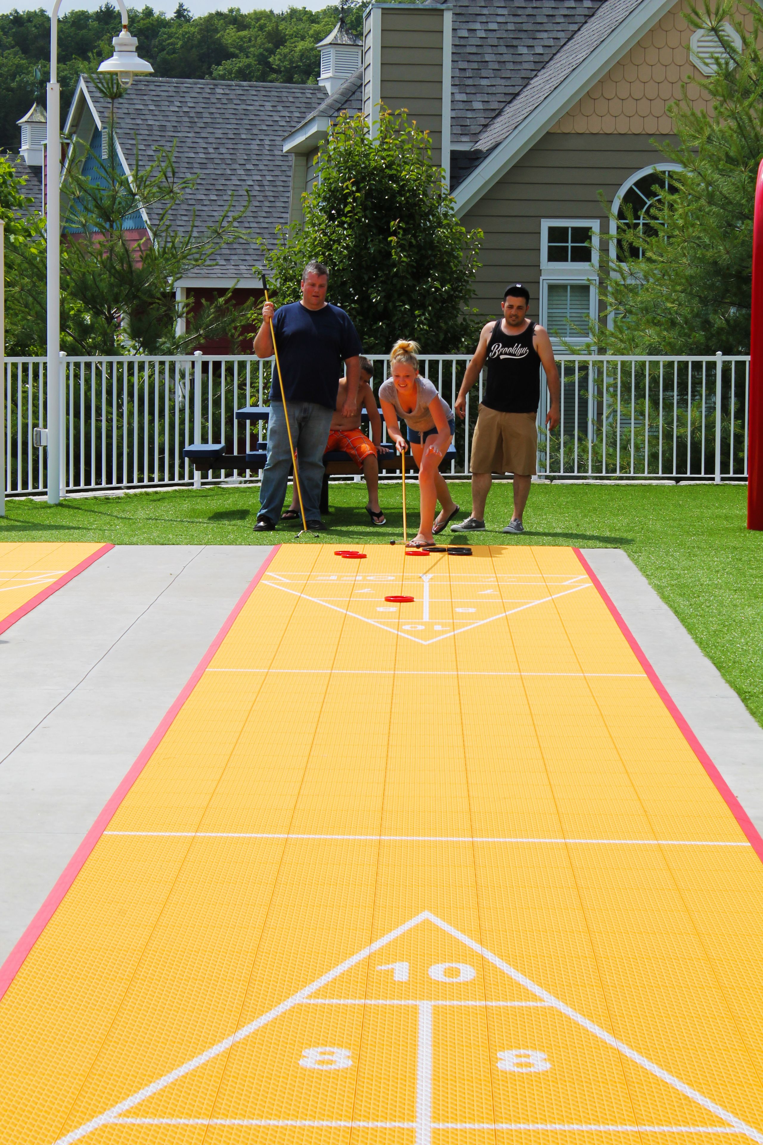 Mesa de shuffleboard com jogadores e uma casa ao fundo.
