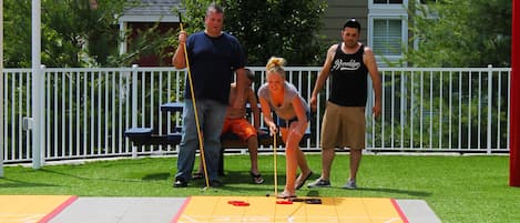 Mesa de shuffleboard com jogadores e uma casa ao fundo.