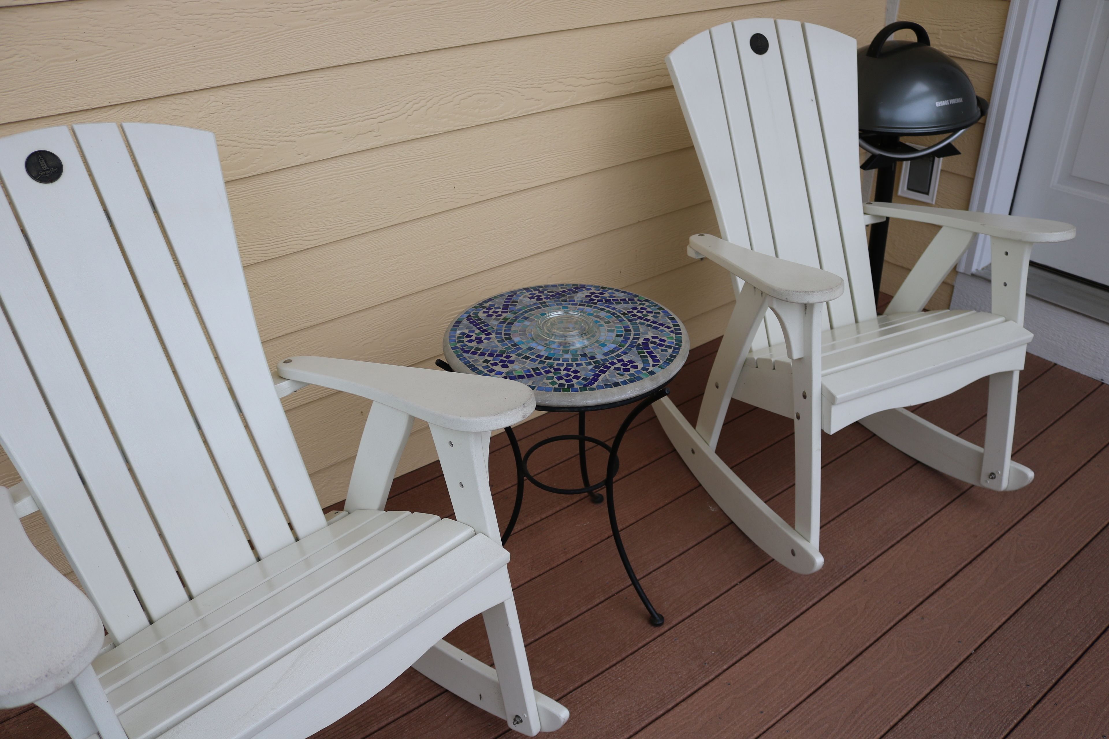 Two white Adirondack chairs and a small round table with a mosaic tile top on a wooden deck.