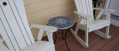 Two white Adirondack chairs and a small round table with a mosaic tile top on a wooden deck.