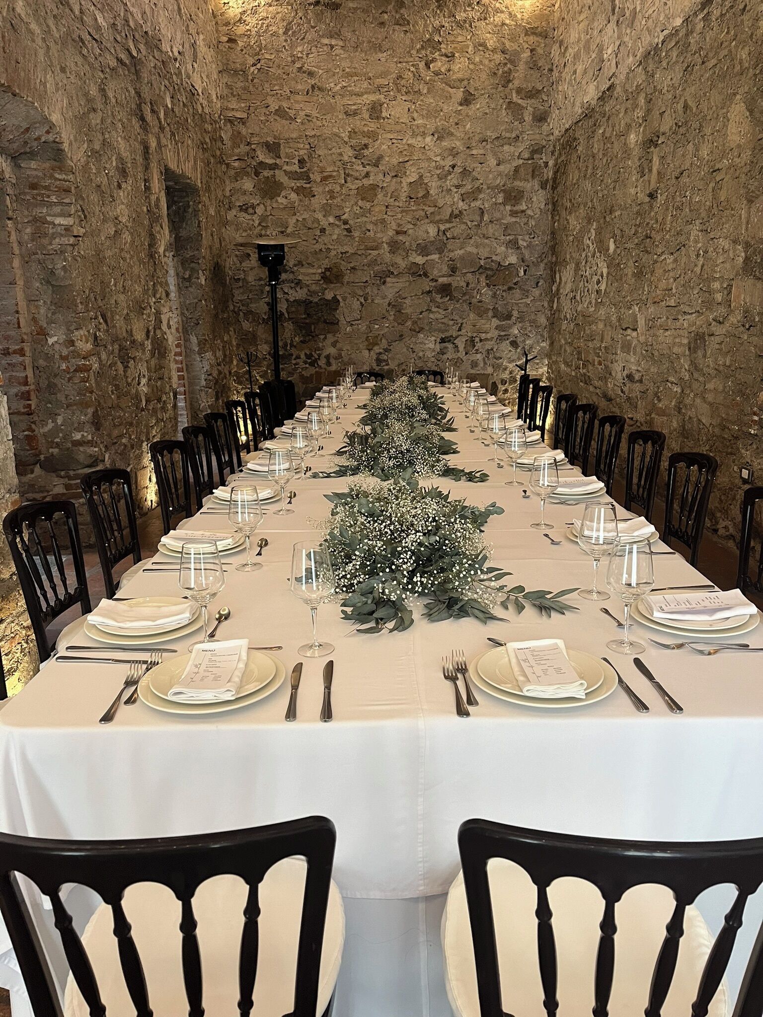 Una larga mesa de comedor con manteles blancos, platos, cubiertos y vasos, flanqueada por sillas negras, en una habitación con paredes de piedra.