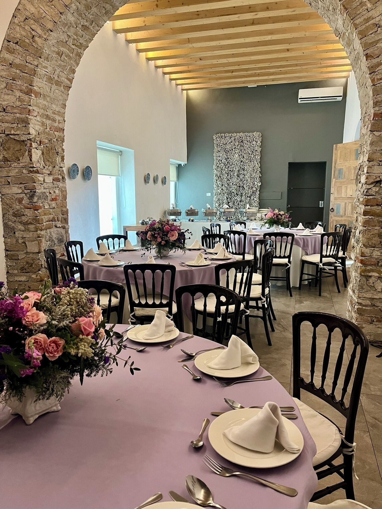 Un comedor con mesas dispuestas para una comida, que presenta un arco de piedra, vigas de madera en el techo y un muro decorativo.