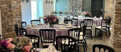 Un comedor con mesas dispuestas para una comida, que presenta un arco de piedra, vigas de madera en el techo y un muro decorativo.