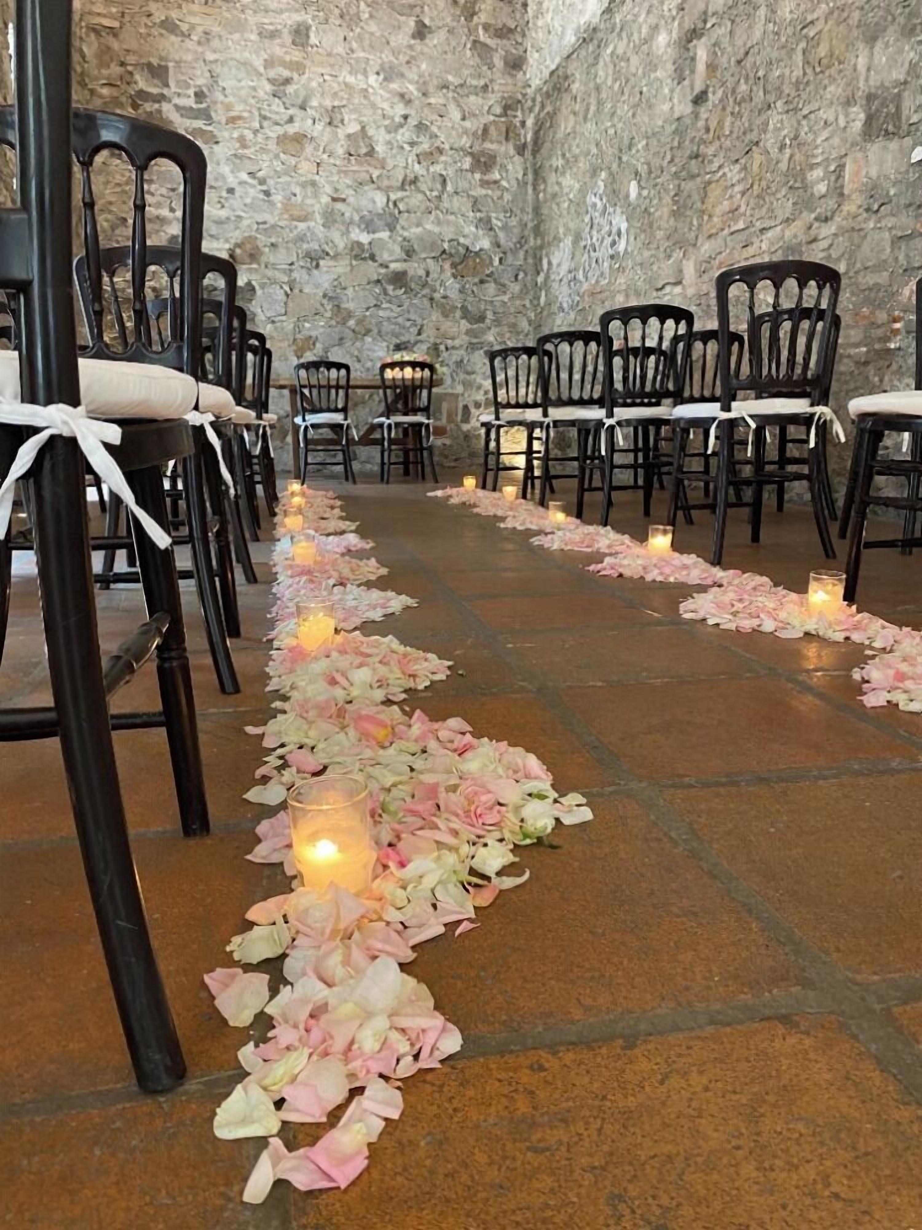 A stone-walled room with rows of black chairs and a rose petal aisle.