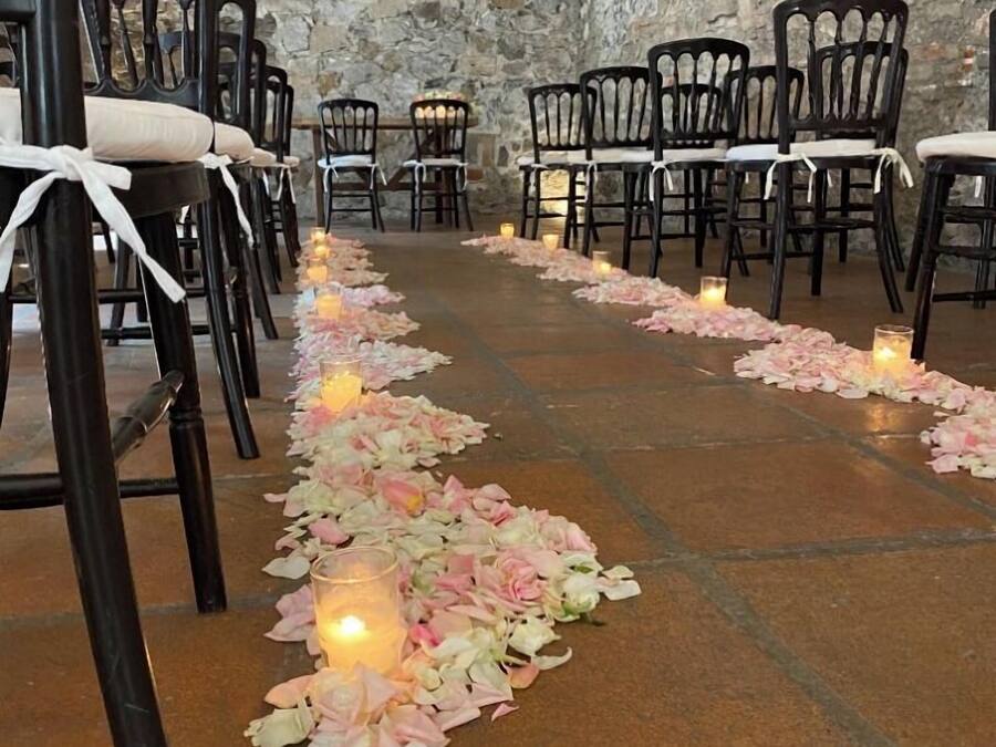 A stone-walled room with rows of black chairs and a rose petal aisle.