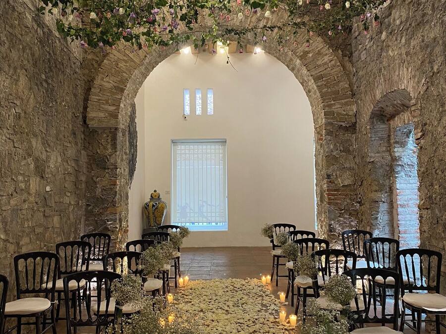 An indoor venue with an arched stone wall, black chairs arranged in rows, a central aisle with flower petals, and hanging floral decorations.