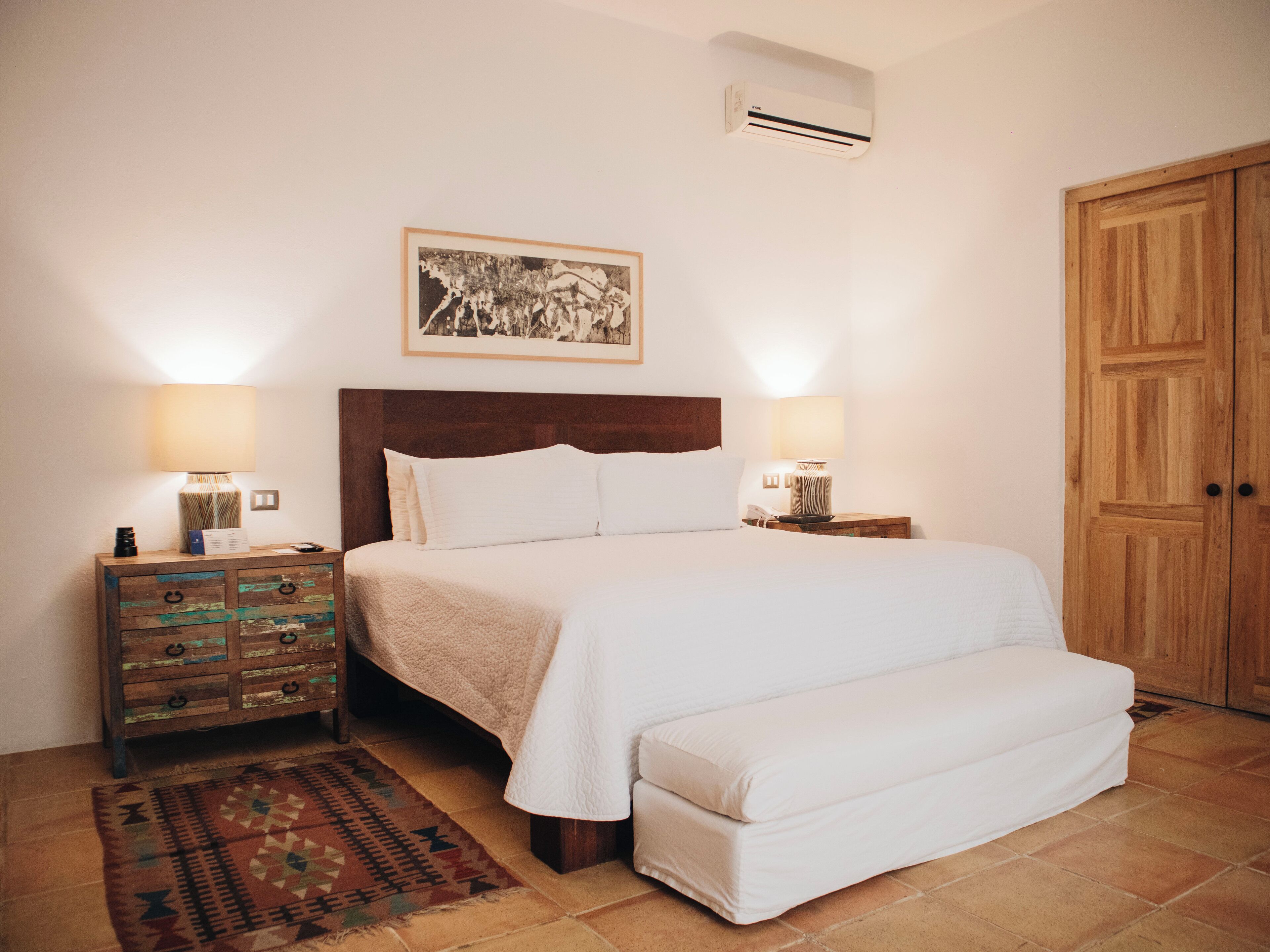 A neatly arranged bedroom with a bed, bedside lamps, a wooden nightstand, a patterned rug, and a wooden wardrobe.