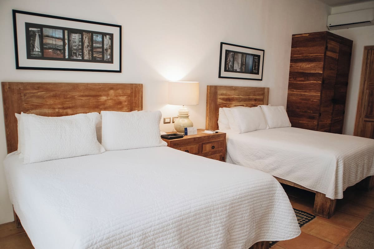Habitación de hotel con dos camas, cabeceros de madera, ropa de cama blanca, una mesita de noche con lámpara y cuadros en la pared.