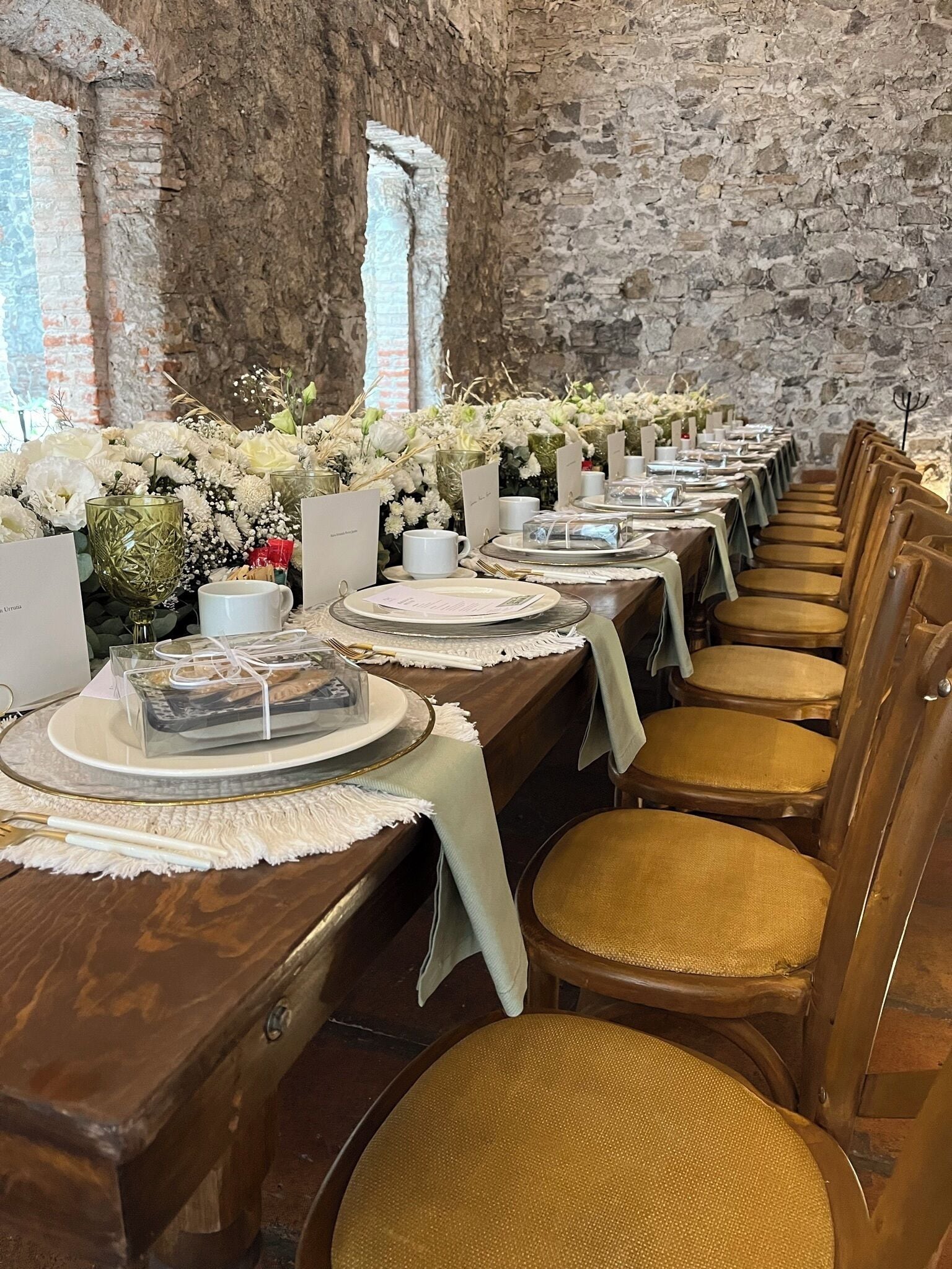 Une longue table à manger, entourée de chaises, et le tout est encadré de murs en pierre et de fenêtres.